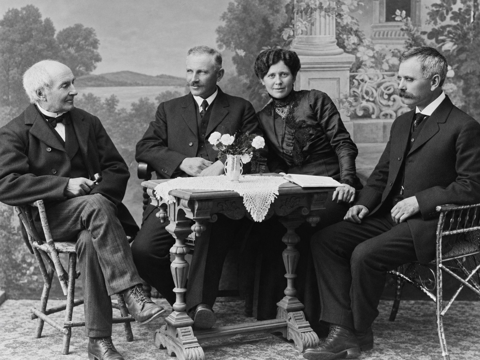 An old white-haired man, two middle-aged men with moustaches and a woman with her hair piled up, seated around a carved table with a vase of flowers, in front of a painted landscape