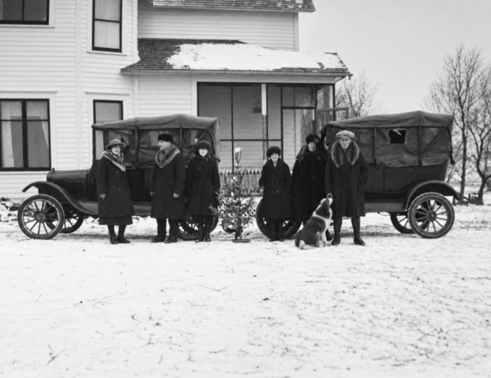 Four women and two men in winter coats, several with fur collars, stand in the snow between two Model T touring cars in front of a white farmhouse, with a small Christmas tree between them and a dog looking up at the young man on the right