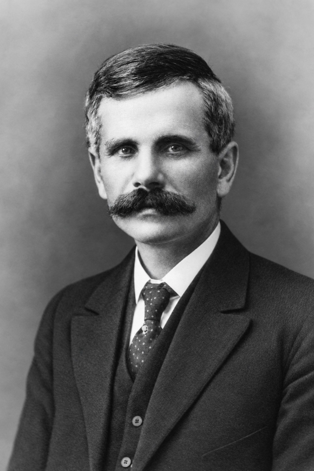 Head and shoulders of a man in his forties with greying hair and a heavy moustache, in a dark suit and patterned tie