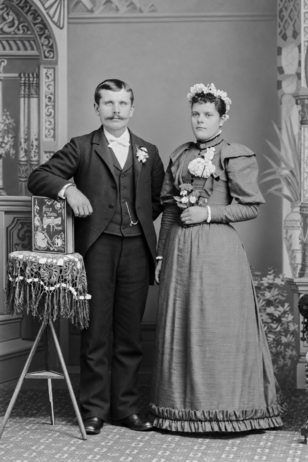 A young man with a large moustache, his arm on a book on a fringed stand, beside a young woman in a long dress with a wreath of flowers in her hair and a bouquet, in front of a painted studio backdrop