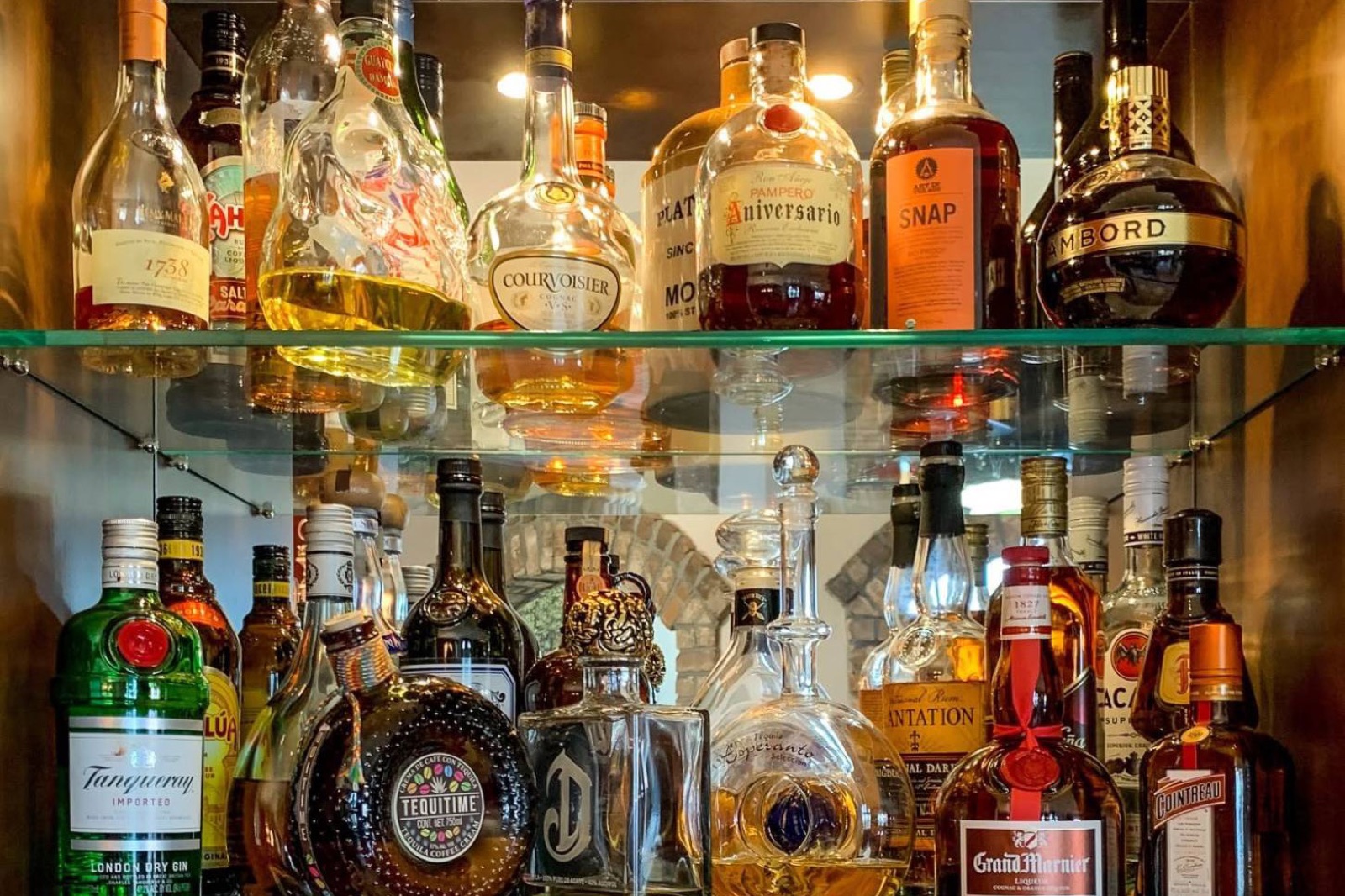 Two lit glass shelves of bottles in the home bar