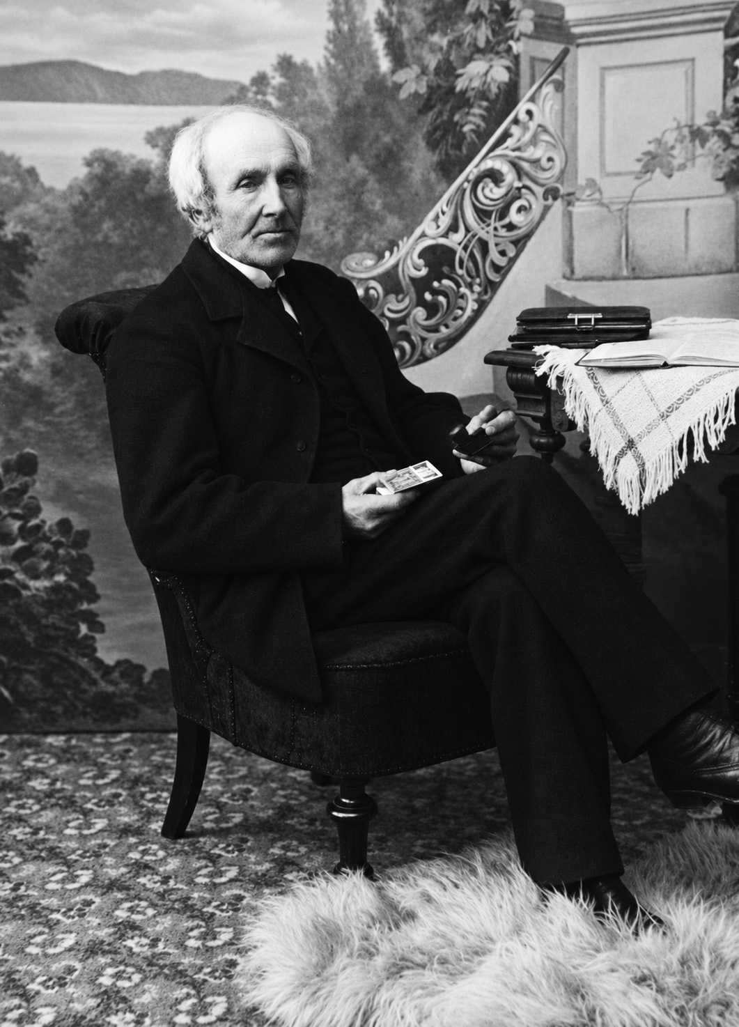 An elderly man with white hair, in a dark suit, seated in a studio before a painted lake scene, holding a small card