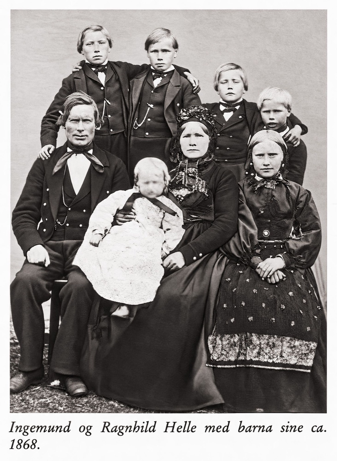 A family portrait: a seated father, mother and teenage girl in Norwegian dress, a small child in white in front of them, and four boys in dark jackets standing behind, with the printed caption Ingemund og Ragnhild Helle med barna sine ca. 1868 below