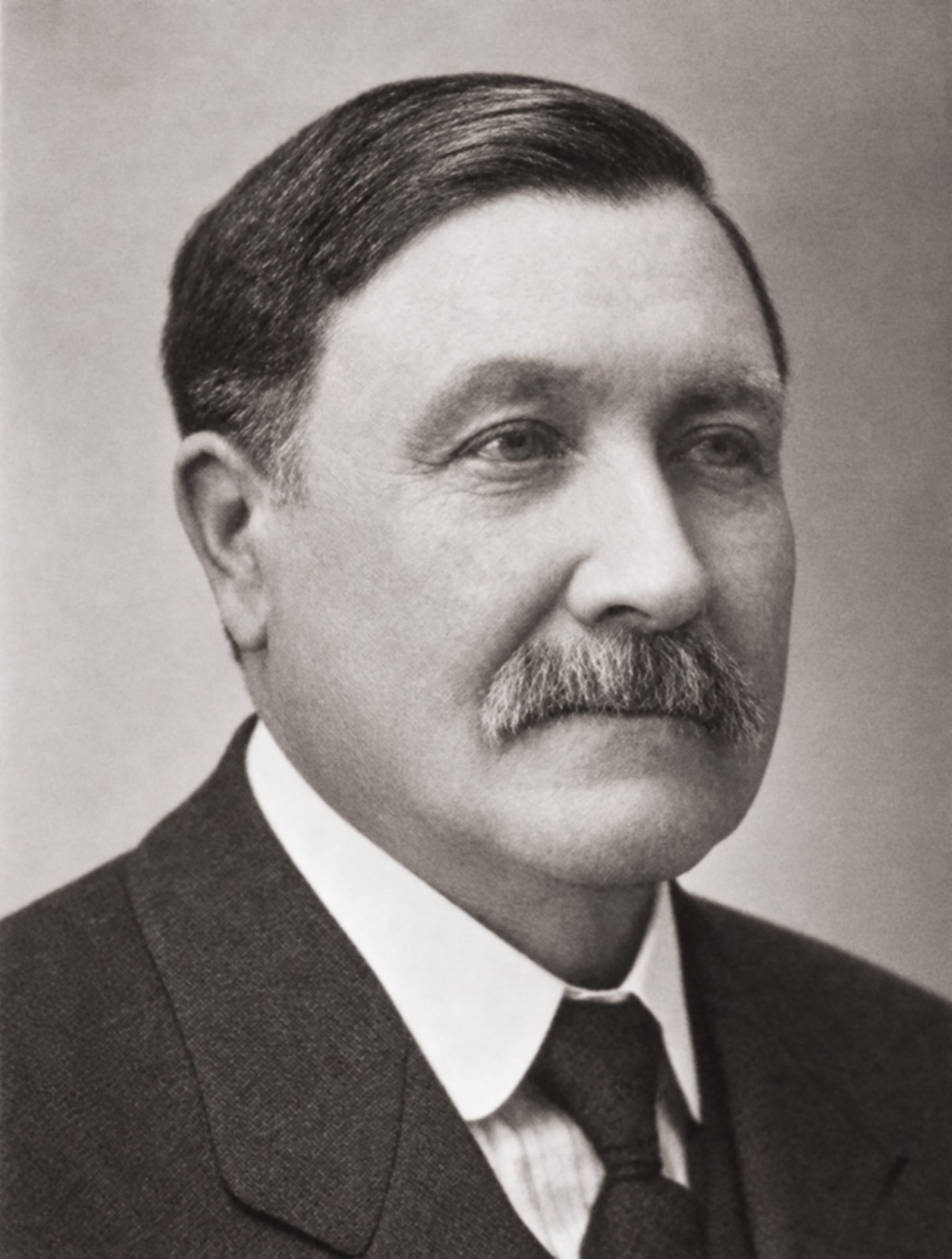 Head and shoulders of a heavyset man with a side part and a grey moustache, in a dark suit, white collar and tie, looking off to the right