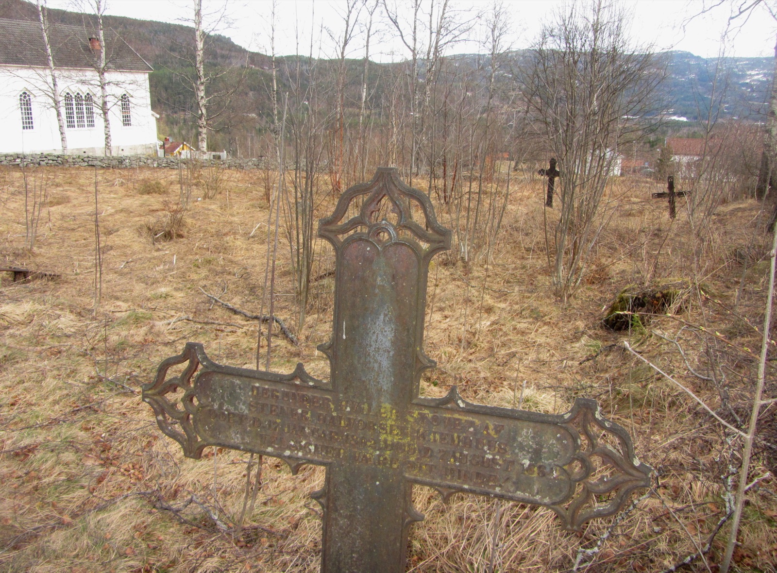 A weathered cast-iron cross with openwork ends and lettered arms, standing in the dead grass of an old churchyard, with a white wooden church and a stone wall behind and two more iron crosses farther off