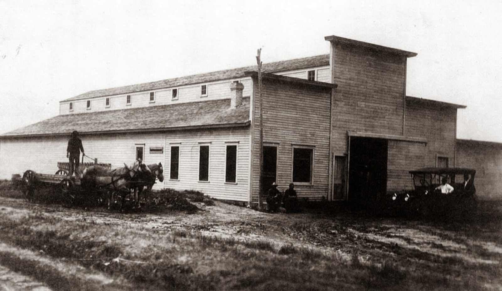 A long two-story lumber shed with a row of small upper windows, a team of horses and a wagon at left, men sitting by the wall, and an early automobile at right