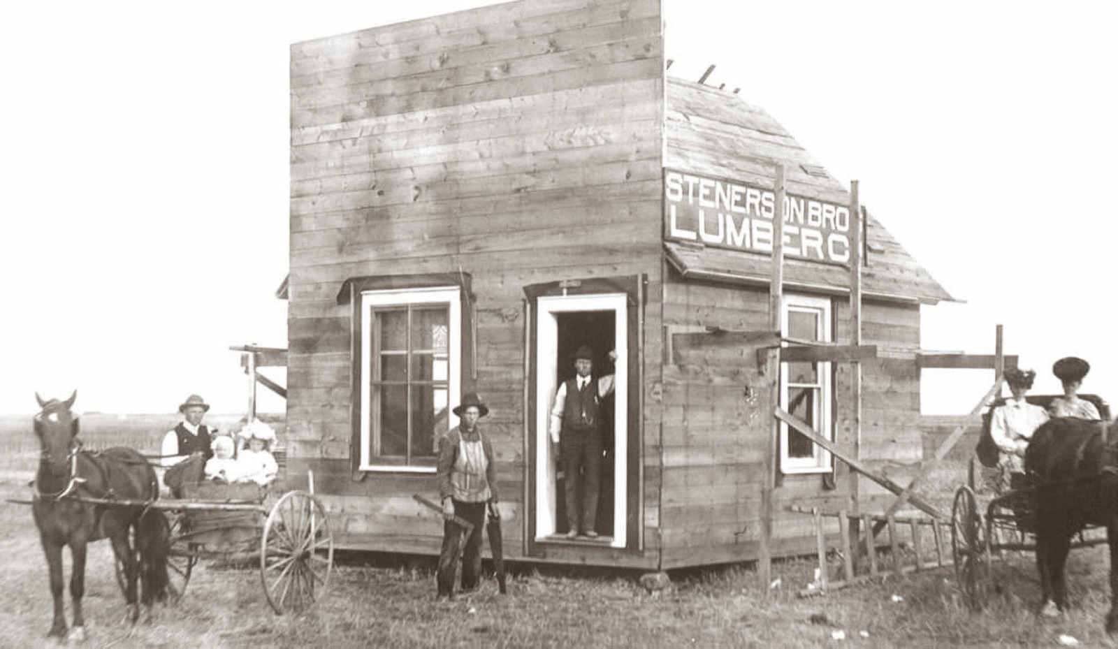 A new frame building with a sign reading STENERSON BRO LUMBER C, ladders and boards against it, a man in the doorway, a boy with a saw in front, and a horse and buggy at each side on open prairie