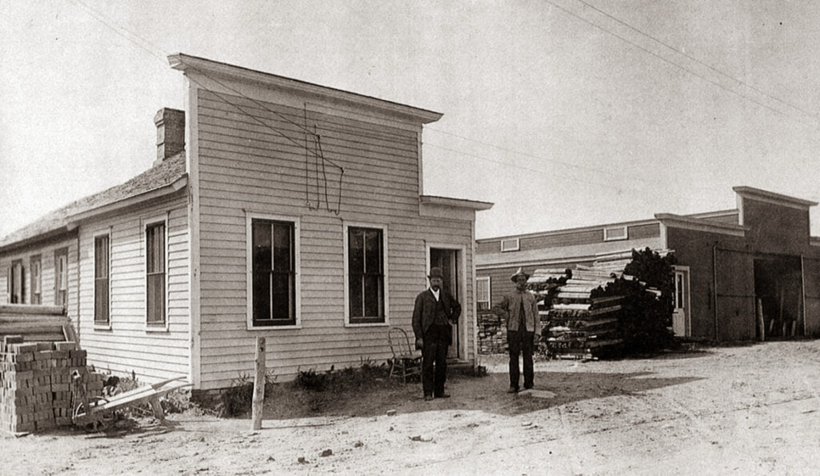 Two men standing outside a small clapboard office with a false front, a stack of bricks and a wheelbarrow at left, and stacked lumber and long sheds behind