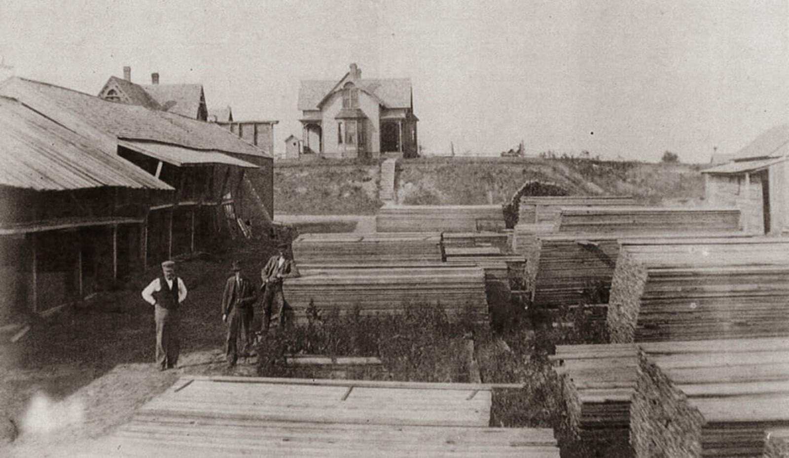 Three men in a lumberyard between long stacks of boards and an open shed, with a large house on the hill beyond