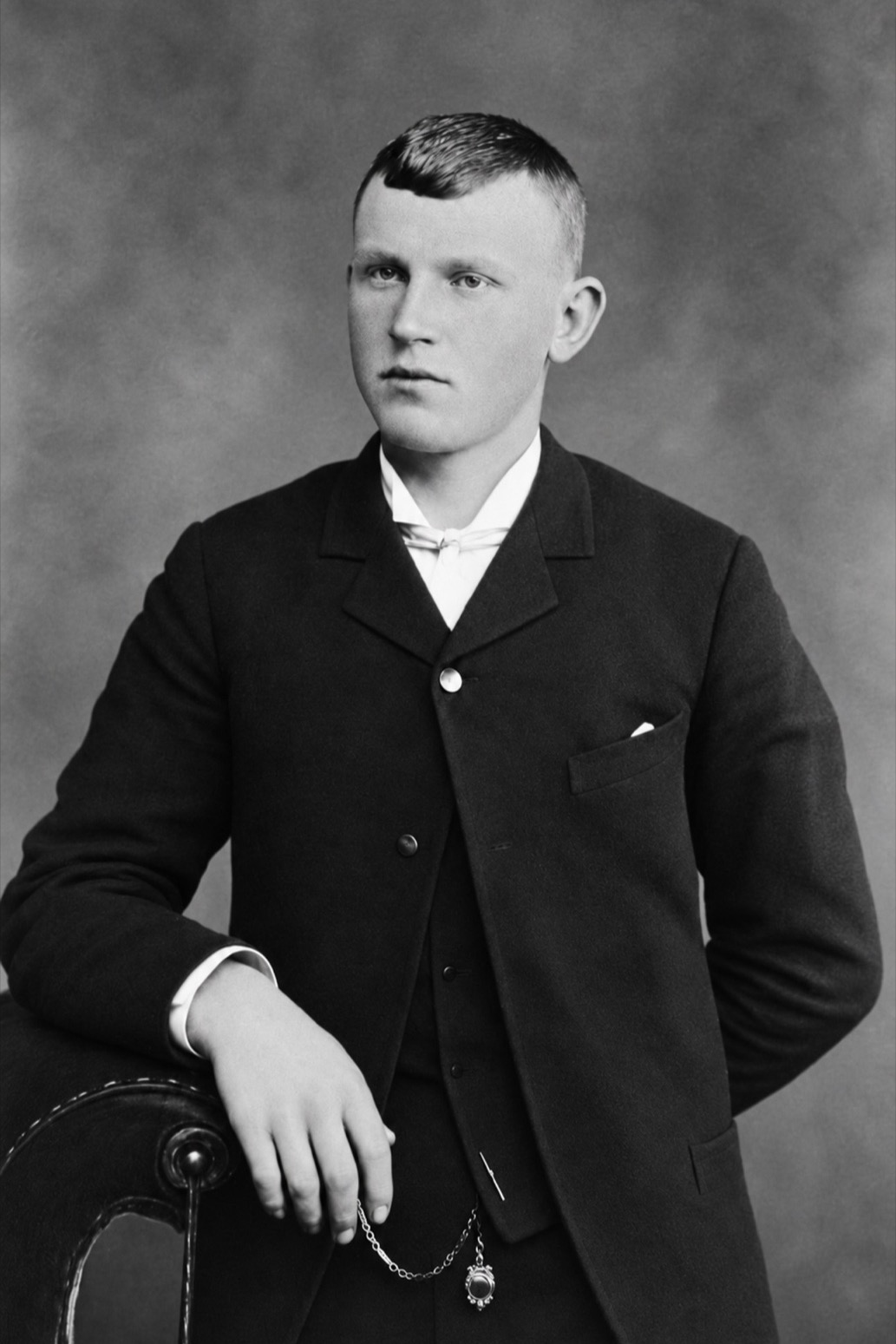 A young man with close-cropped hair in a dark suit and watch chain, one hand resting on the back of a chair