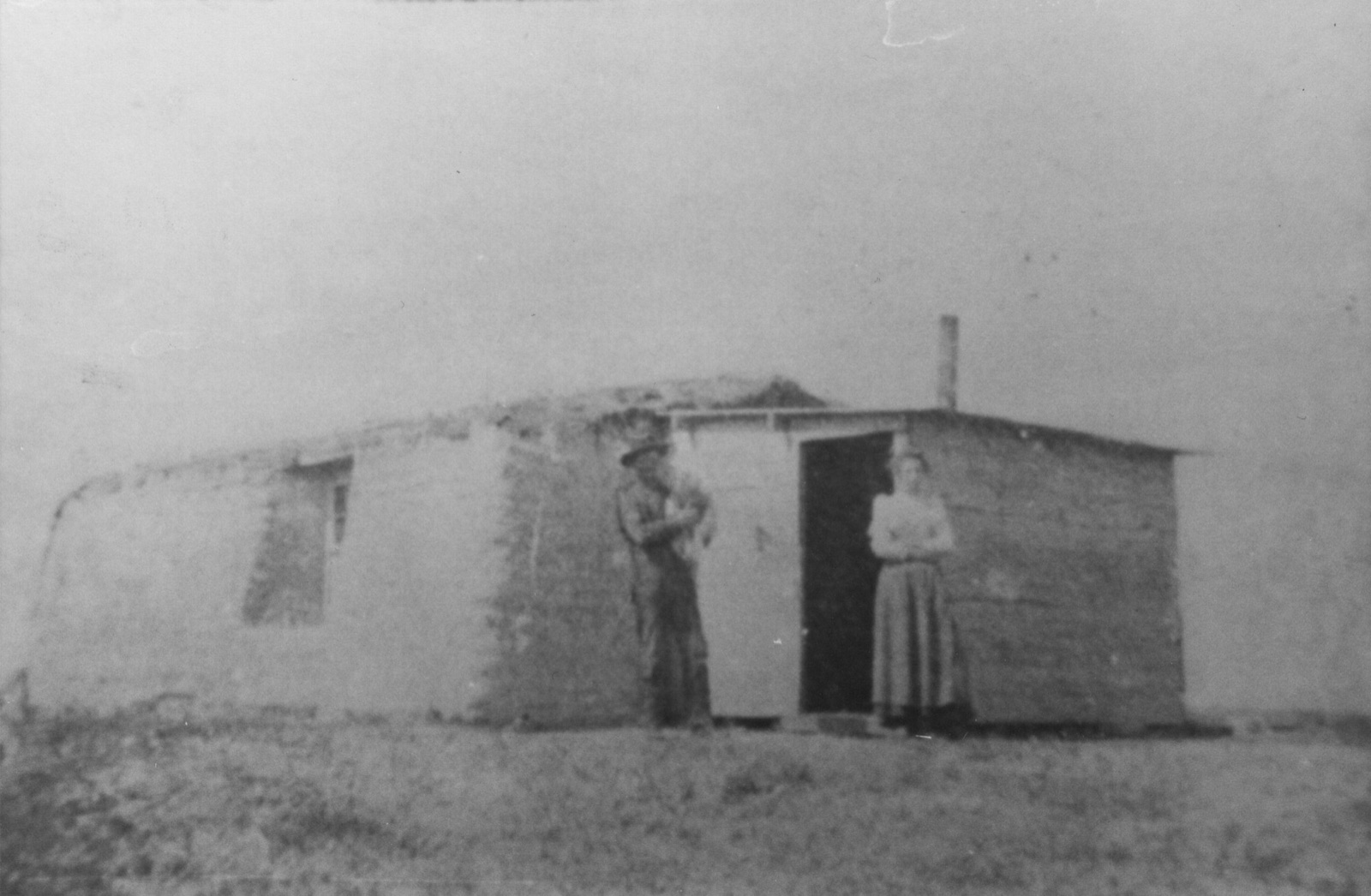 A sod house with a board-built end and a stovepipe, on open prairie; a man in a hat and a woman in a long skirt stand either side of the open door